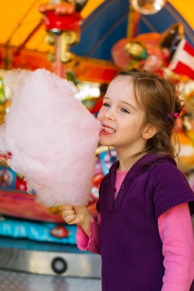 Ein kleines Mädchen isst rosa Zuckerwatte vor einem bunten Kirmeskarussell.