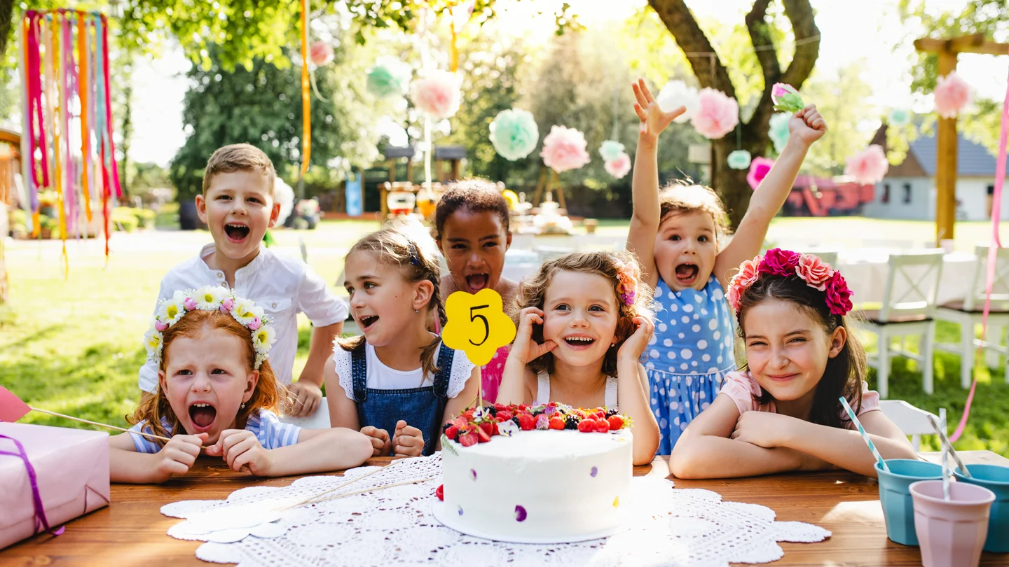 Sechs Kinder mit Blumenkränzen im Haar jubeln an einem festlich gedeckten Geburtstagstisch mit Torte im Garten.